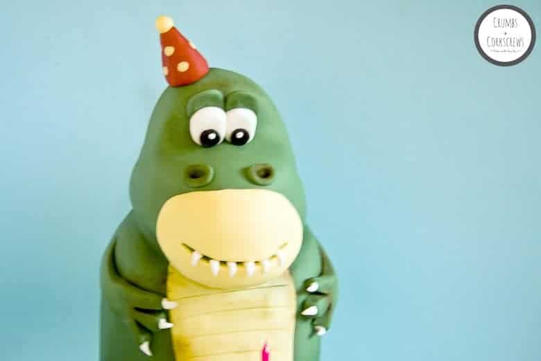 Cute green T-Rex dinosaur cake wearing a red party hat