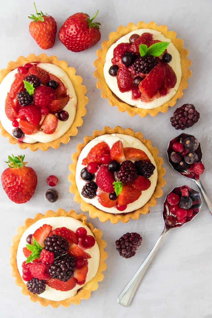 Top down view of pastry tarts, filled with cream and summer fruits. Scattered strawberries and blackcurrants on the table.