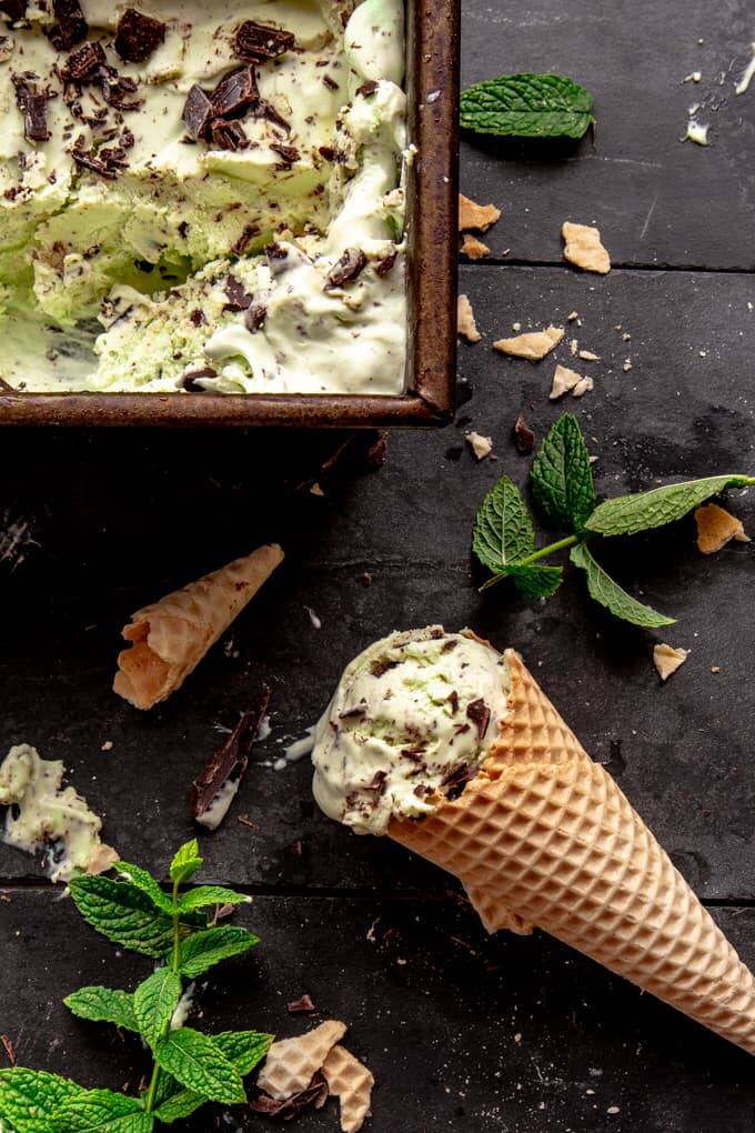 Mint ice cream in a waffle cone on a black slate with chocolate pieces and mint leaves
