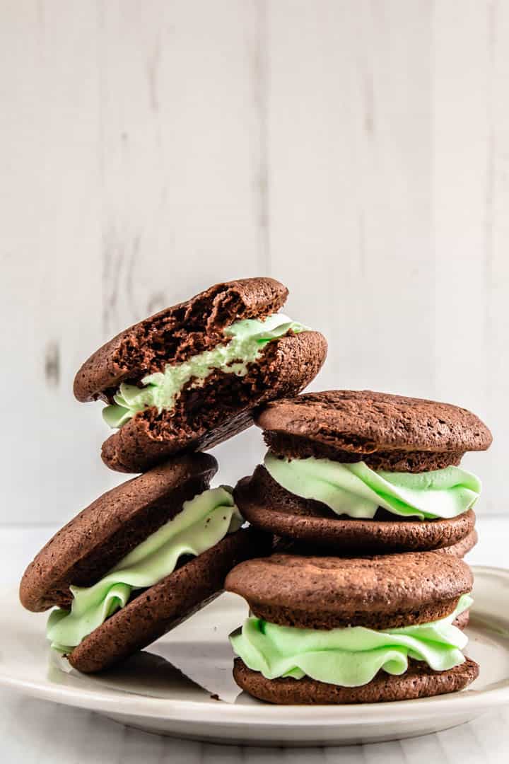 Four chocolate whoopie pies filled with green mint frosting, piled on top of each other on a plate. One has a bite taken out of it.