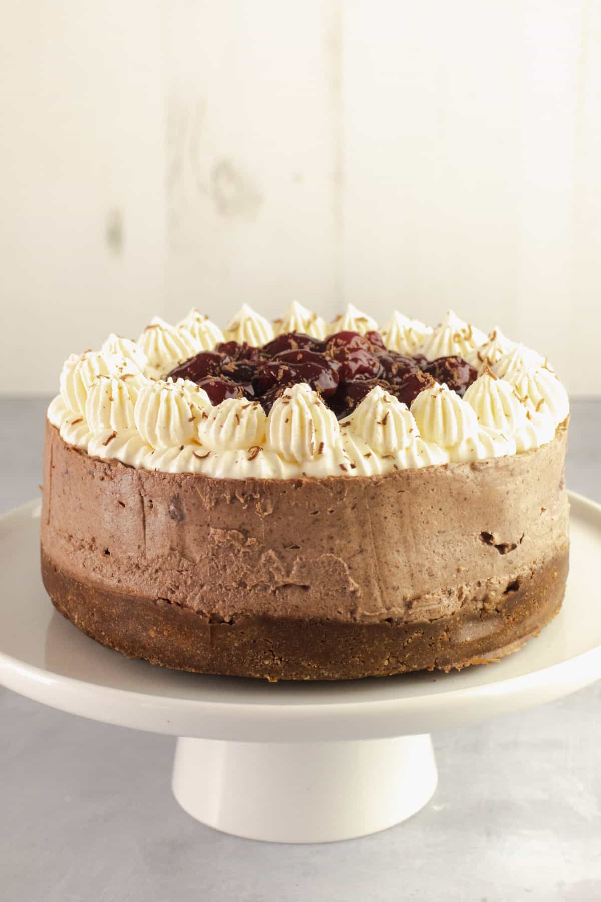 Chocolate cheesecake on a white cake stand with whipped cream topping and black cherries