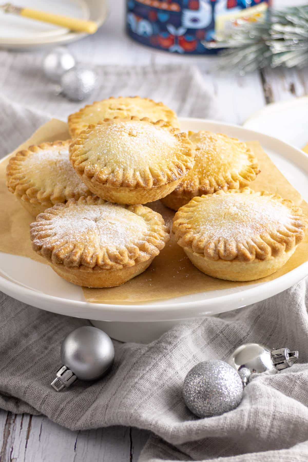 A pile of sweet mince pies on a white stand surrounded by silver baubles