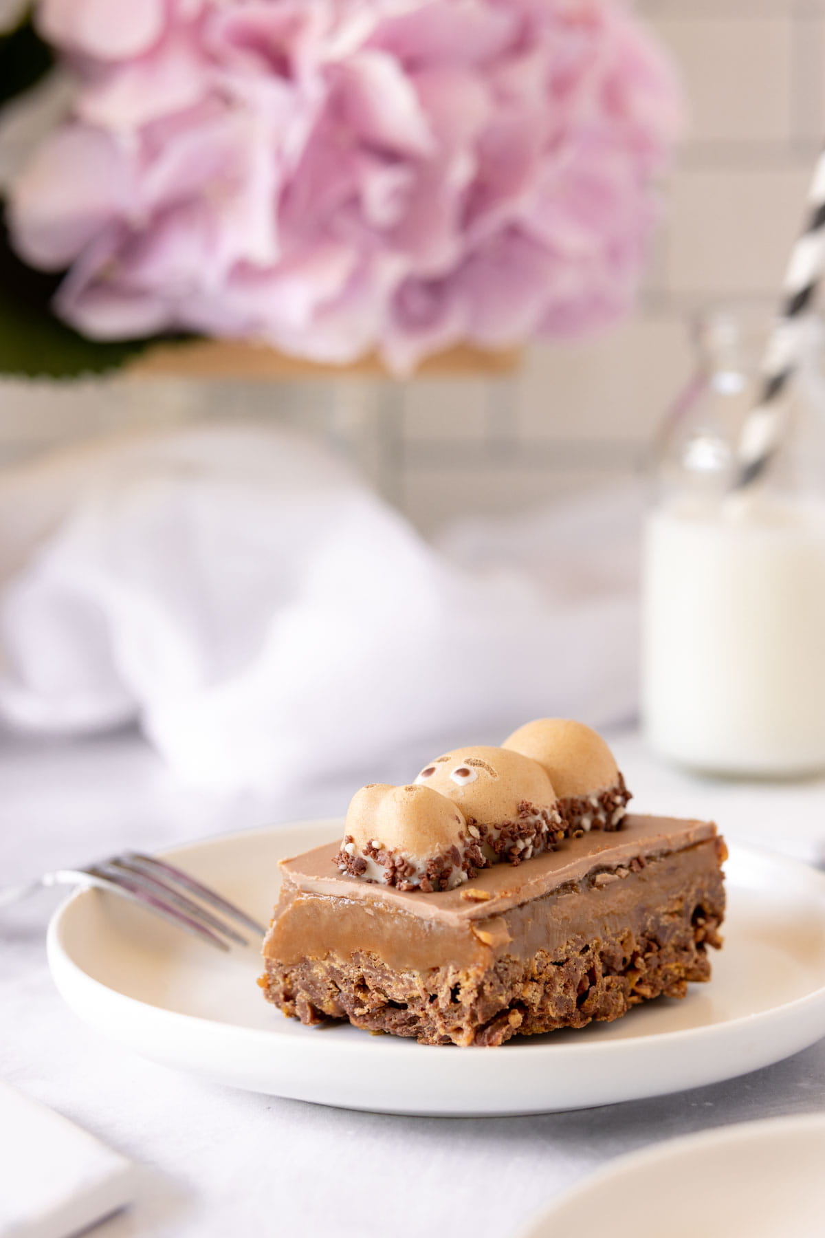 Chocolate cornflake slice with a layer of caramel sauce on a whit plate with a pink hydrangea flower