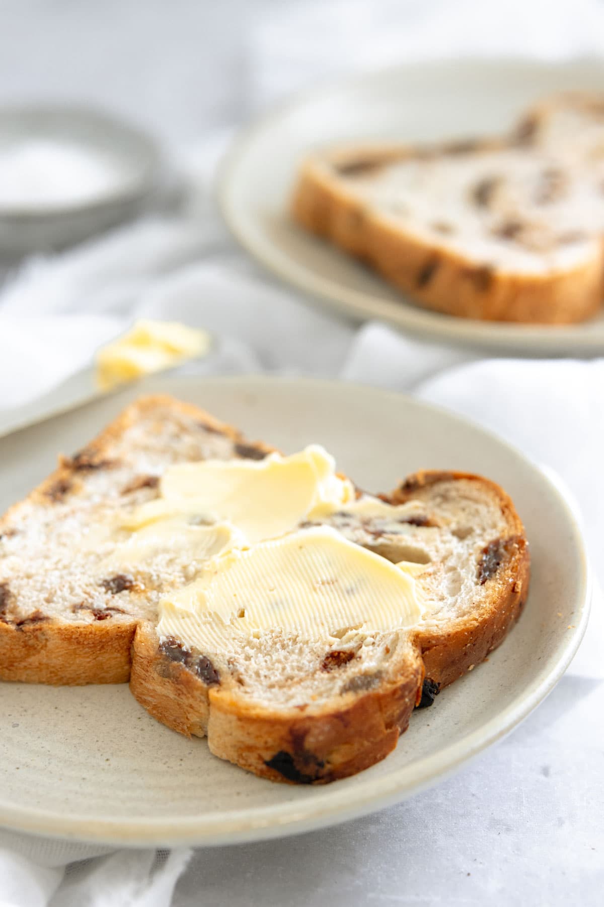 A slice of fruit loaf on a plate with a butter knife, spread with salted butter