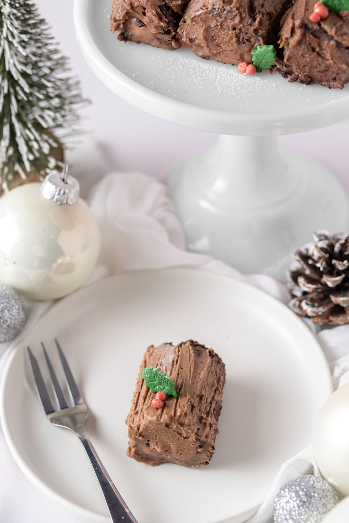 Mini chocolate yule log on a plate with a fork next to a pine cone and silver bauble