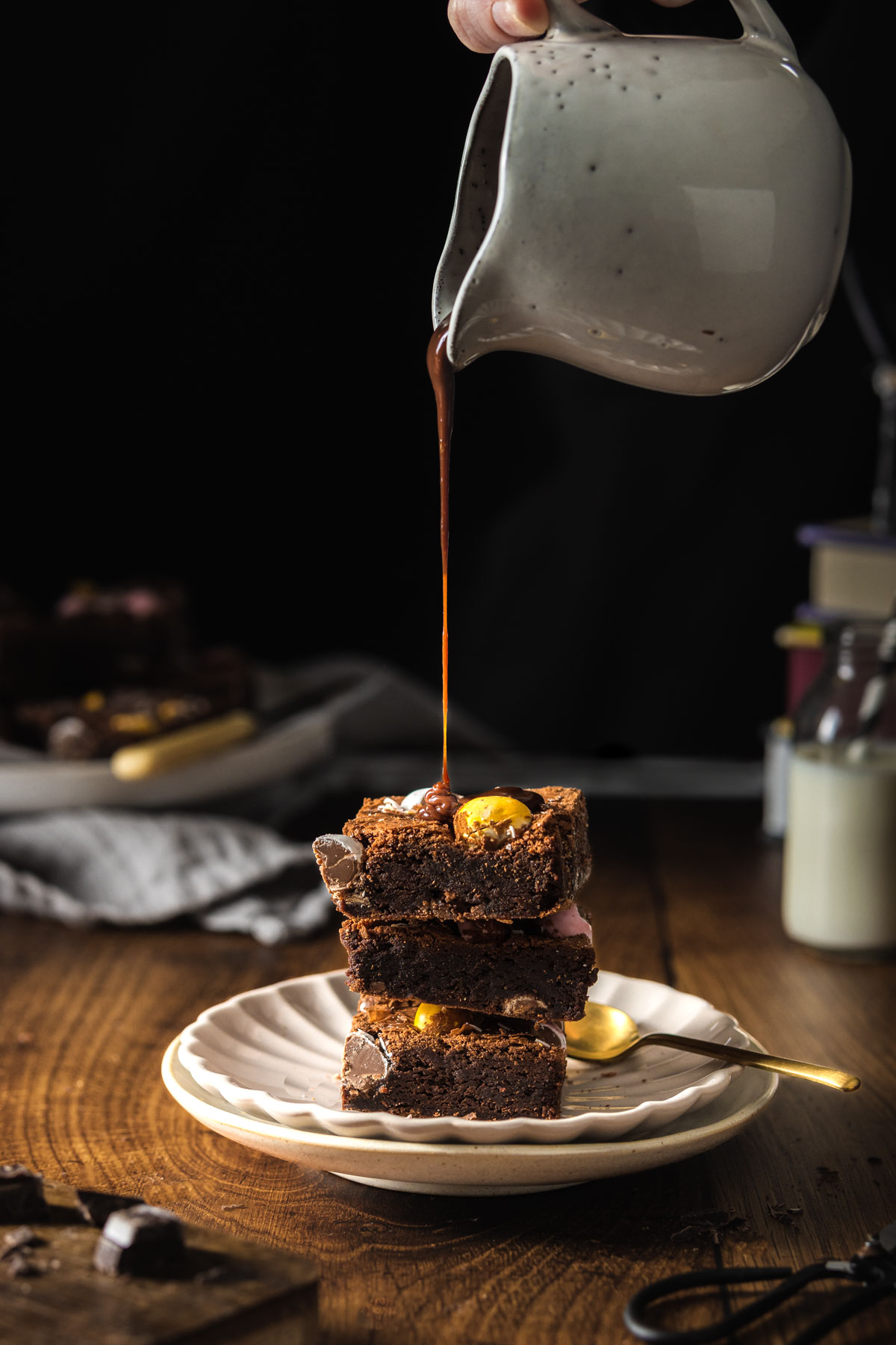 Jug of chocolate sauce being poured over a stack of three chocolate brownies filled with Mini Eggs