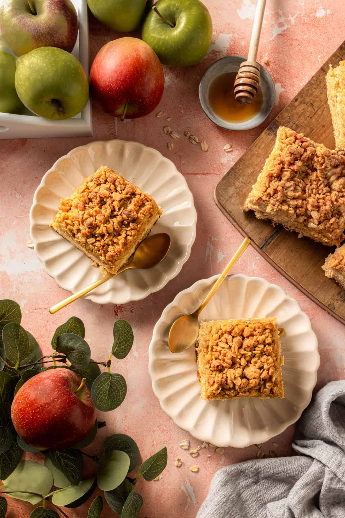 Two slices of apple crumble tray bake cake on plates, surrounded by apples