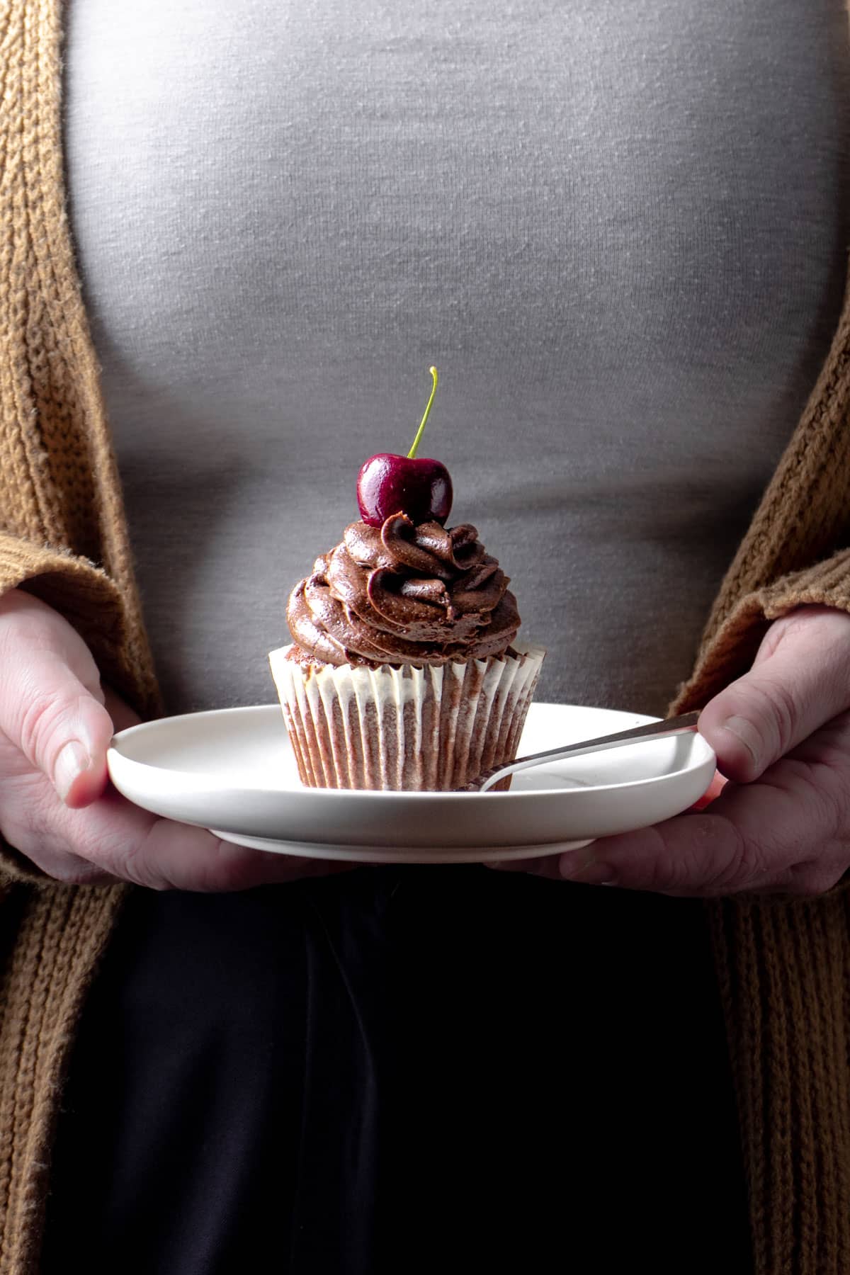 A chocolate cupcake with a cherry on the top being held on a plate - Photography and styling by Lou Carruthers, Food Photographer