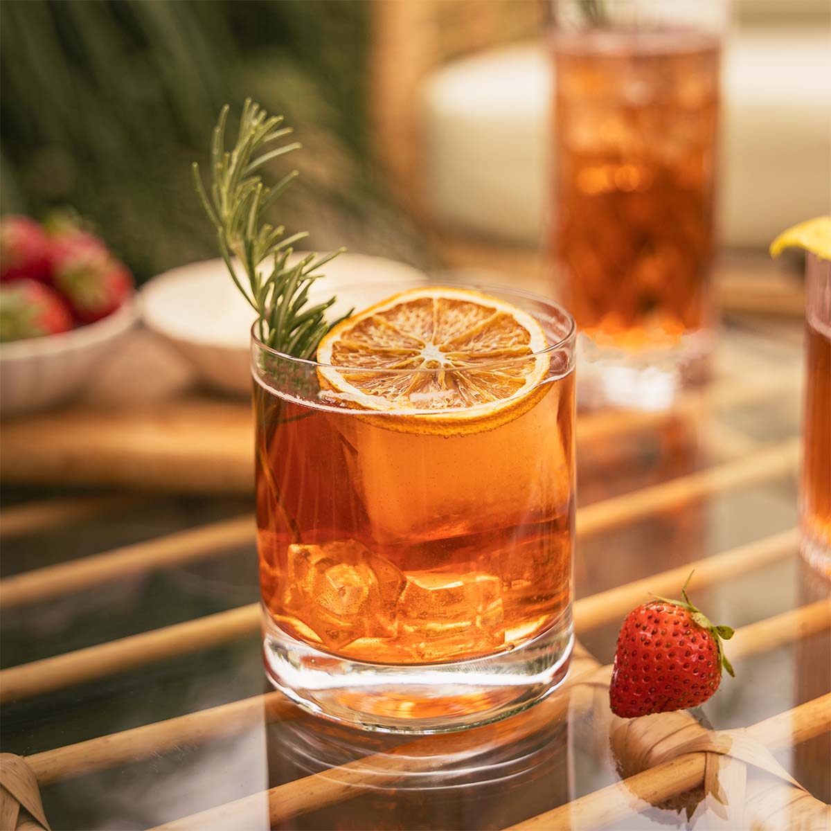 Close-up of a summer cocktail in a short glass, garnished with a slice of dried orange and fresh rosemary, with strawberries and cream in the background.