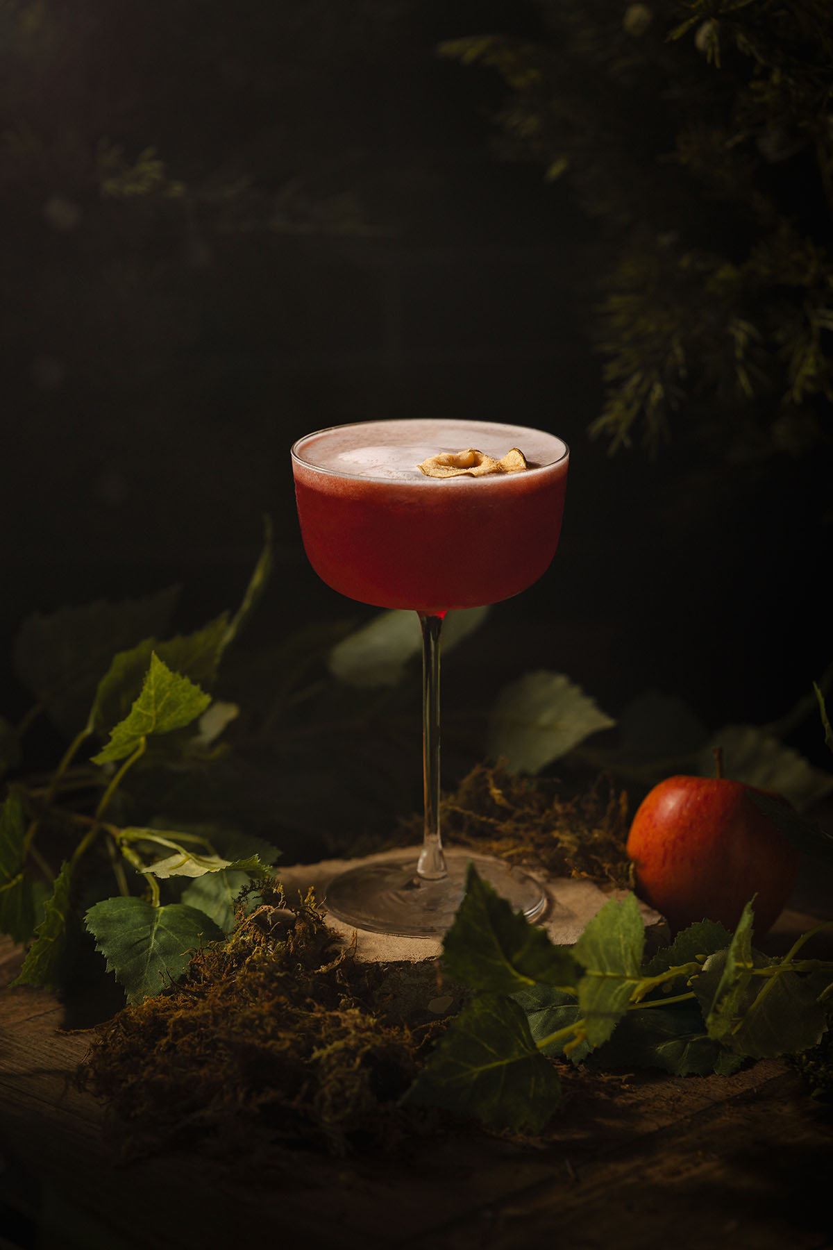 Alt Text: A rich red mocktail topped with delicate apple foam and garnish, photographed by Lou Carruthers. The image captures a cinematic woodland mood with soft shadows, green foliage, and a single apple hinting at temptation.