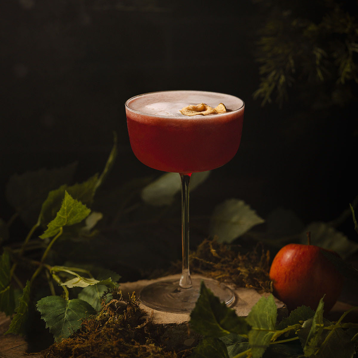 A rich red mocktail topped with delicate apple foam and garnish, photographed by Lou Carruthers. The image captures a cinematic woodland mood with soft shadows, green foliage, and a single apple hinting at temptation.