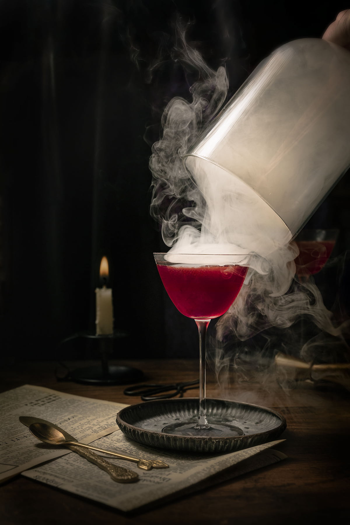 A dark magenta gin and rum cocktail in a coupe glass as cherry smoke curls dramatically around it while the cloche is lifted, captured by Lou Carruthers in moody, candlelit tones.