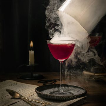A dark magenta gin and rum cocktail in a coupe glass as cherry smoke curls dramatically around it while the cloche is lifted, captured by Lou Carruthers in moody, candlelit tones.
