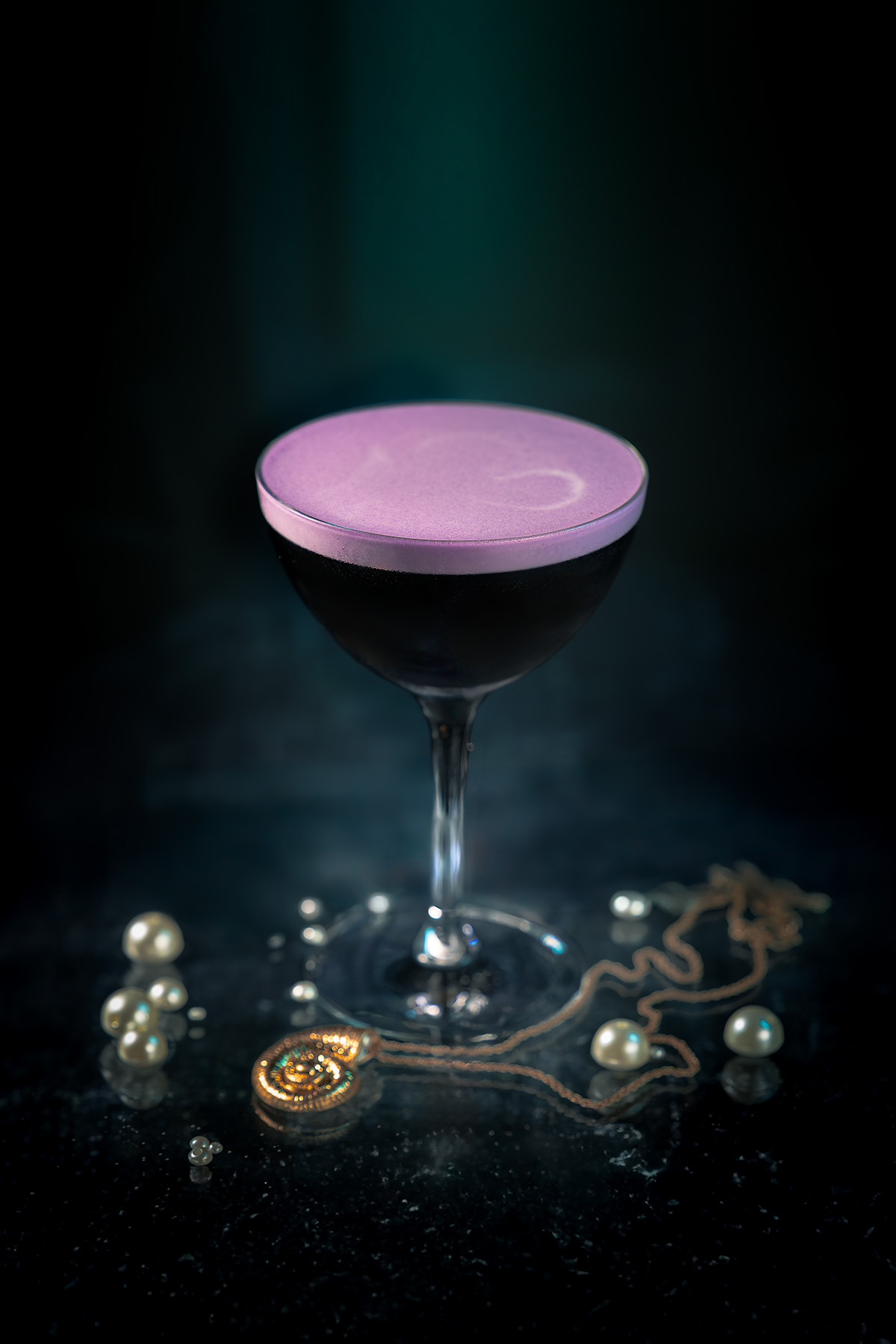 A moody portrait of the Ursula cocktail by Lou Carruthers, styled with pearls and a golden seashell necklace on a dark ocean-like surface with teal light beams above.