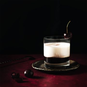 A black-and-white Cruella de Vil cocktail with a dark cherry garnish, captured by Lou Carruthers on a moody red background with soft spotlighting.
