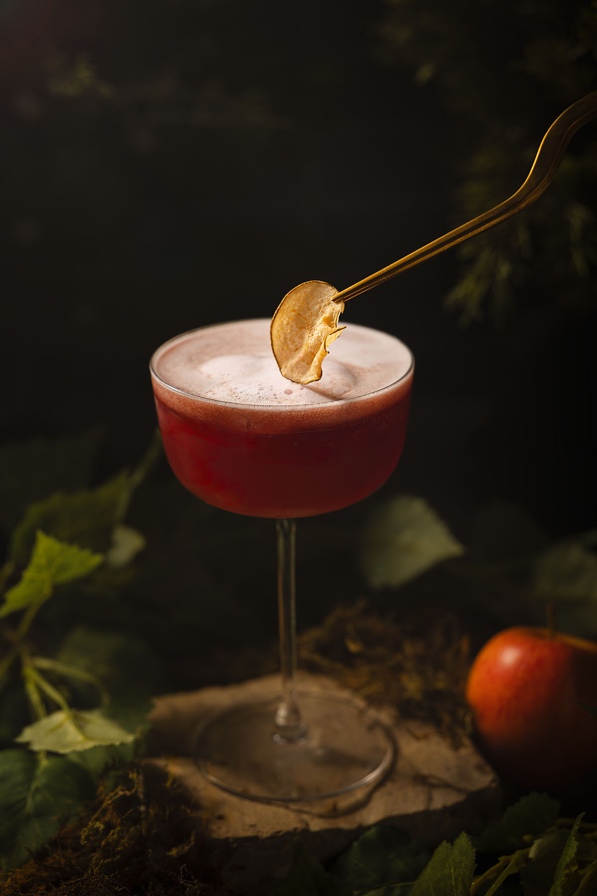 A red apple mocktail in a coupe glass, caught under golden light as a dried apple slice is lowered delicately into the foam. Styled by Lou Carruthers with moss, leaves, and woodland textures inspired by the Snow White fairy tale.
