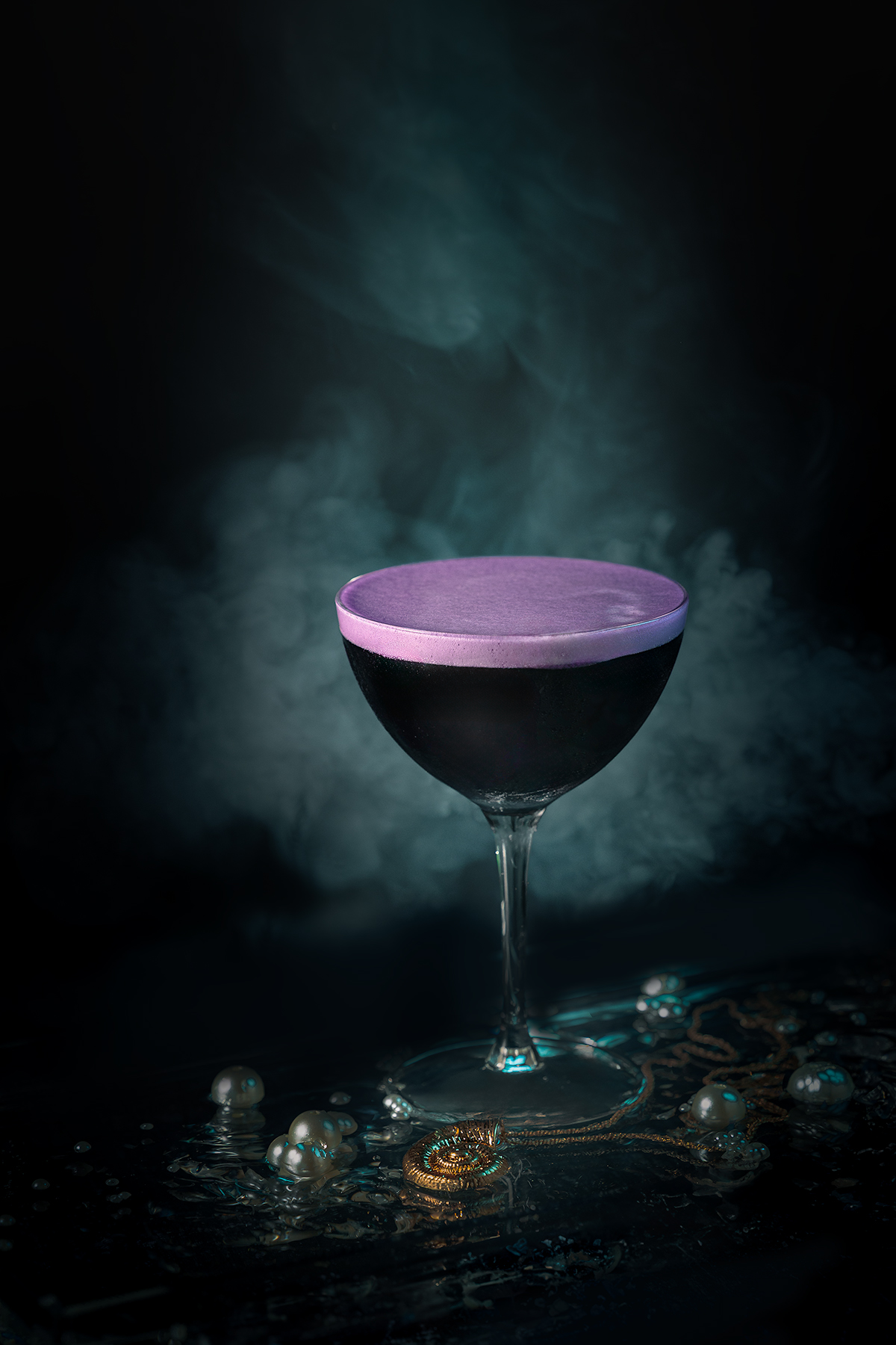 A black coupe cocktail with violet foam surrounded by mist and pearls, photographed by Lou Carruthers to evoke Ursula’s undersea lair and her spellbinding magic.