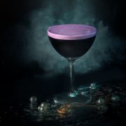 A black coupe cocktail with violet foam surrounded by mist and pearls, photographed by Lou Carruthers to evoke Ursula’s undersea lair and her spellbinding magic.
