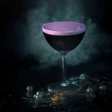 A black coupe cocktail with violet foam surrounded by mist and pearls, photographed by Lou Carruthers to evoke Ursula’s undersea lair and her spellbinding magic.