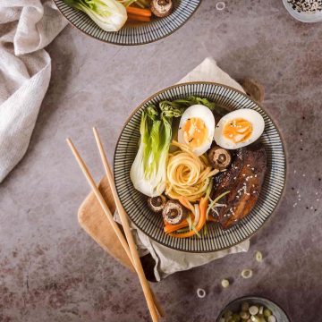Flatlay of a ramen bowl with caramel pork belly, vegetables, noodles, and soft-boiled egg halves, styled for Crumbs & Corkscrews by Lou Carruthers.