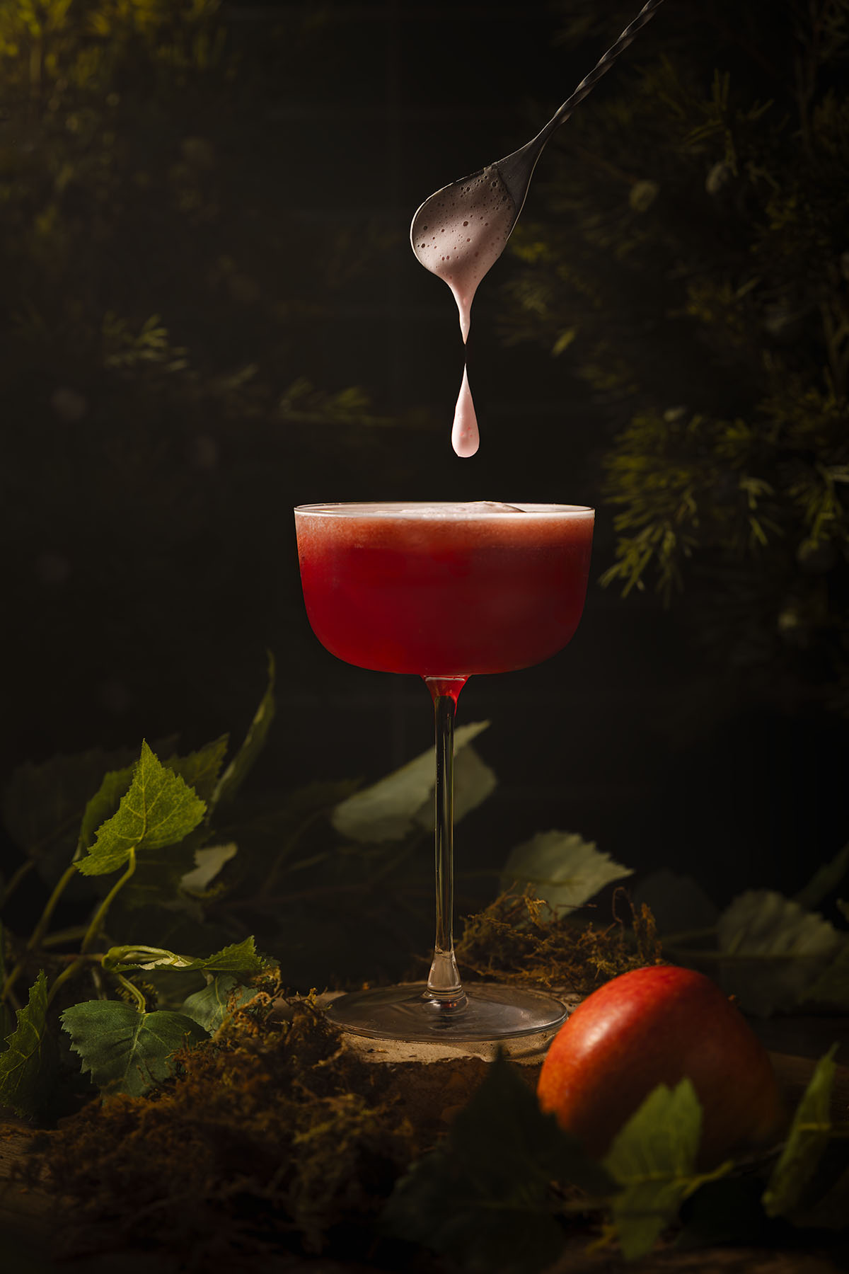 A cinematic cocktail photograph by Lou Carruthers featuring a deep red mocktail inspired by the Evil Queen. Foam drips from a silver spoon above a coupe glass surrounded by moss, leaves, and a single red apple, evoking dark woodland magic.