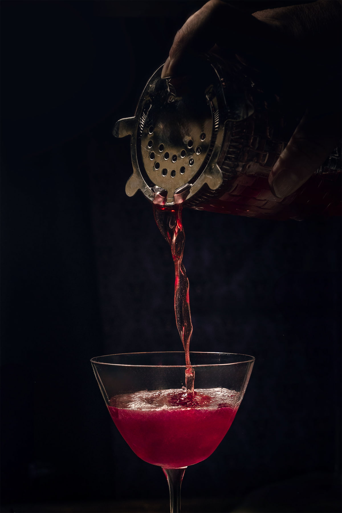 A close-up shot of the magenta Maleficent cocktail as it pours silkily from a shaker into a coupe glass, glowing with deep red light, photographed by Lou Carruthers.