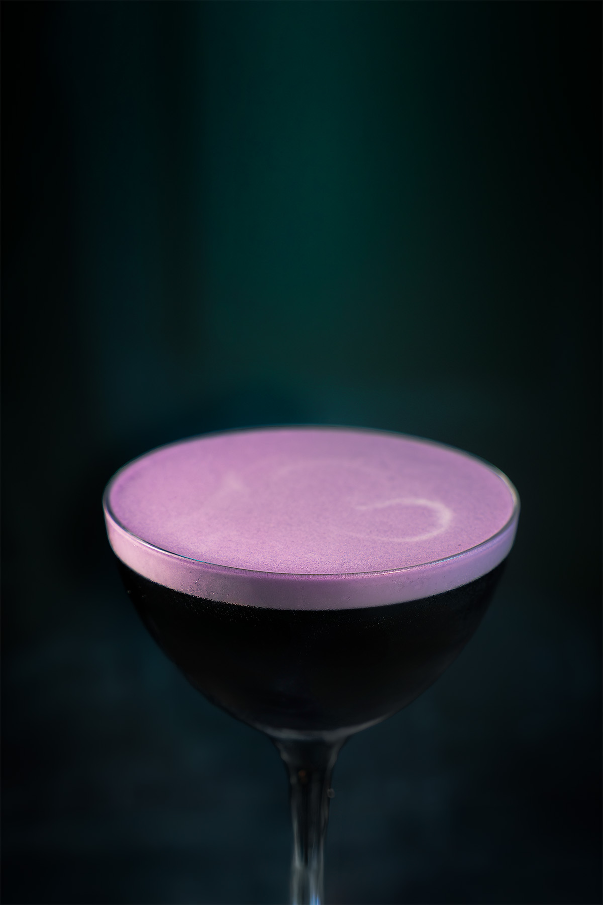 A cinematic dark cocktail inspired by Disney’s Ursula, shot by Lou Carruthers. A deep black drink with soft lilac foam under teal light, capturing the calm tension of the ocean depths.