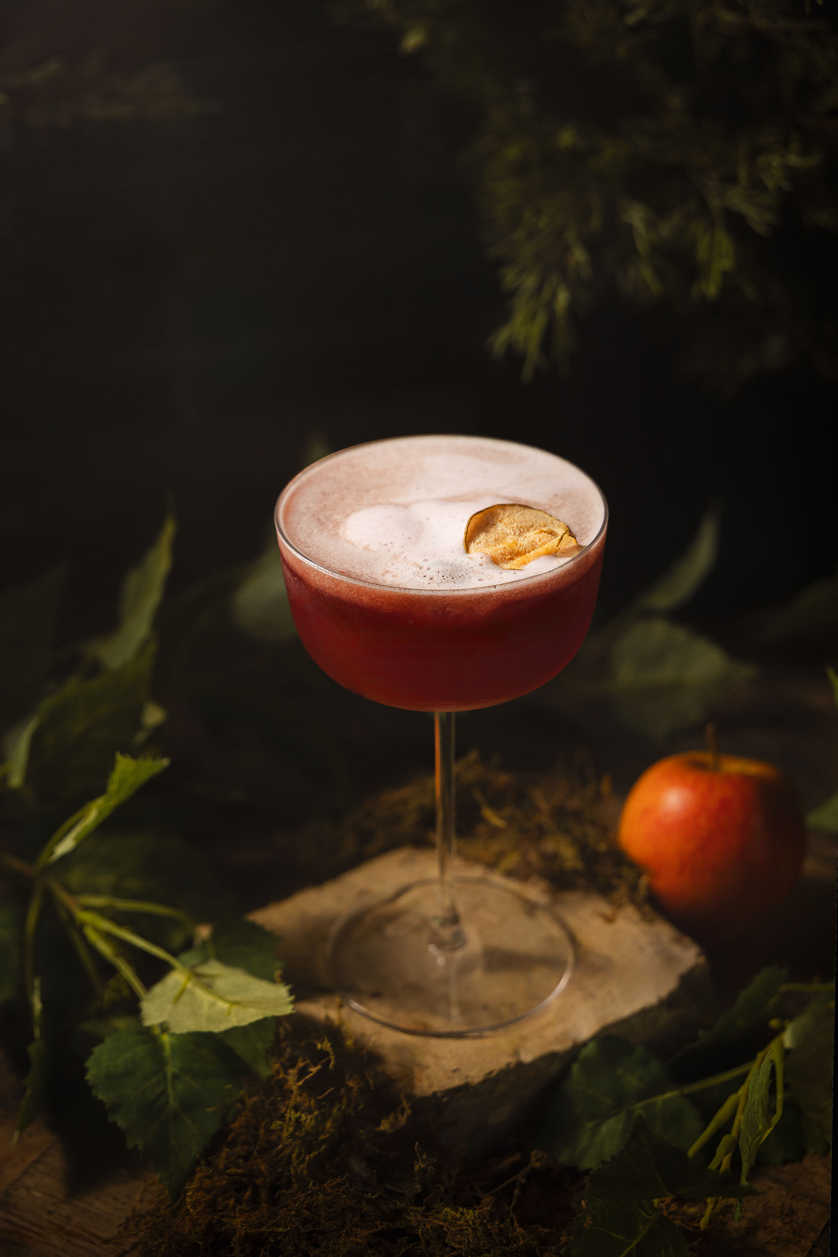 A rich red mocktail topped with delicate apple foam and garnish, photographed by Lou Carruthers. The image captures a cinematic woodland mood with soft shadows, green foliage, and a single apple hinting at temptation.