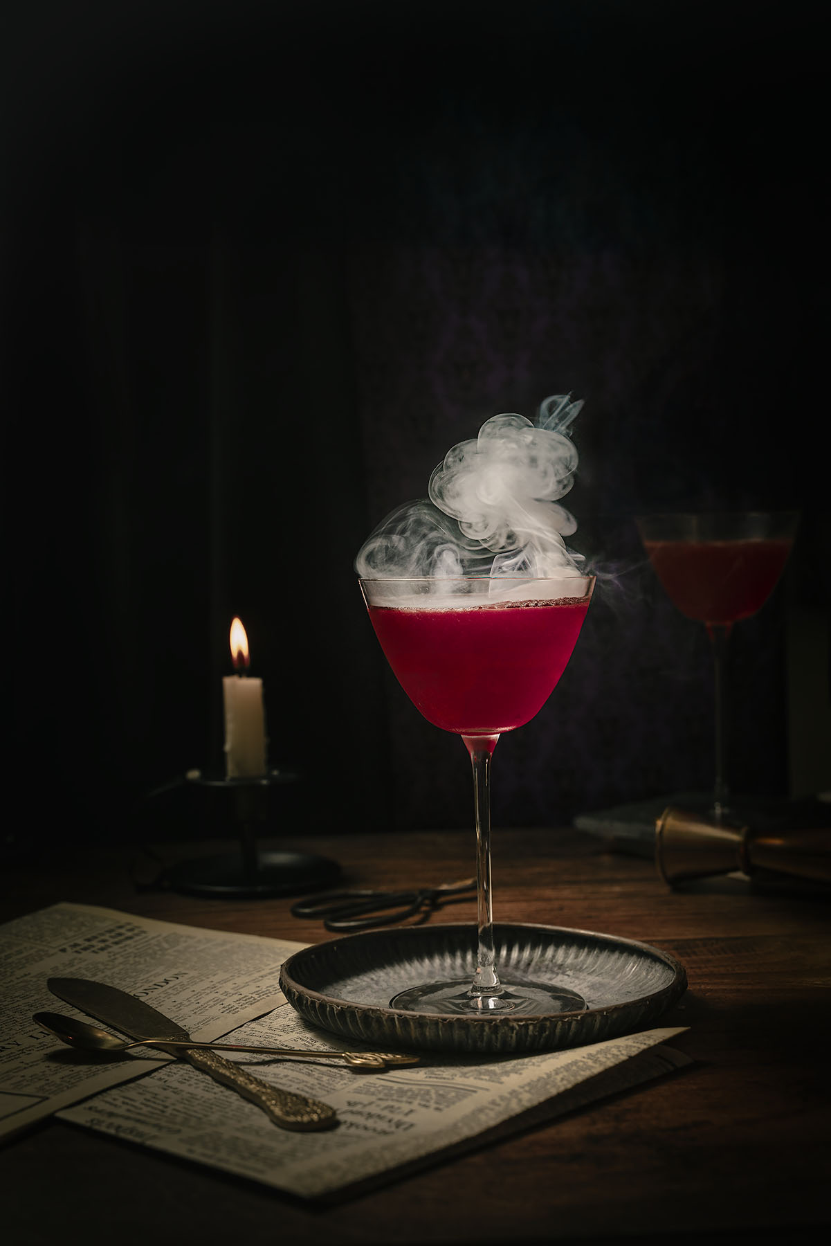 The Maleficent cocktail by Lou Carruthers, styled in a candlelit Halloween scene with vintage cutlery, old newspaper pages, and a smoky magenta gin drink. The moody lighting and swirling vapour capture a darkly elegant, villain-inspired atmosphere — perfect for Halloween cocktails and seasonal party ideas.