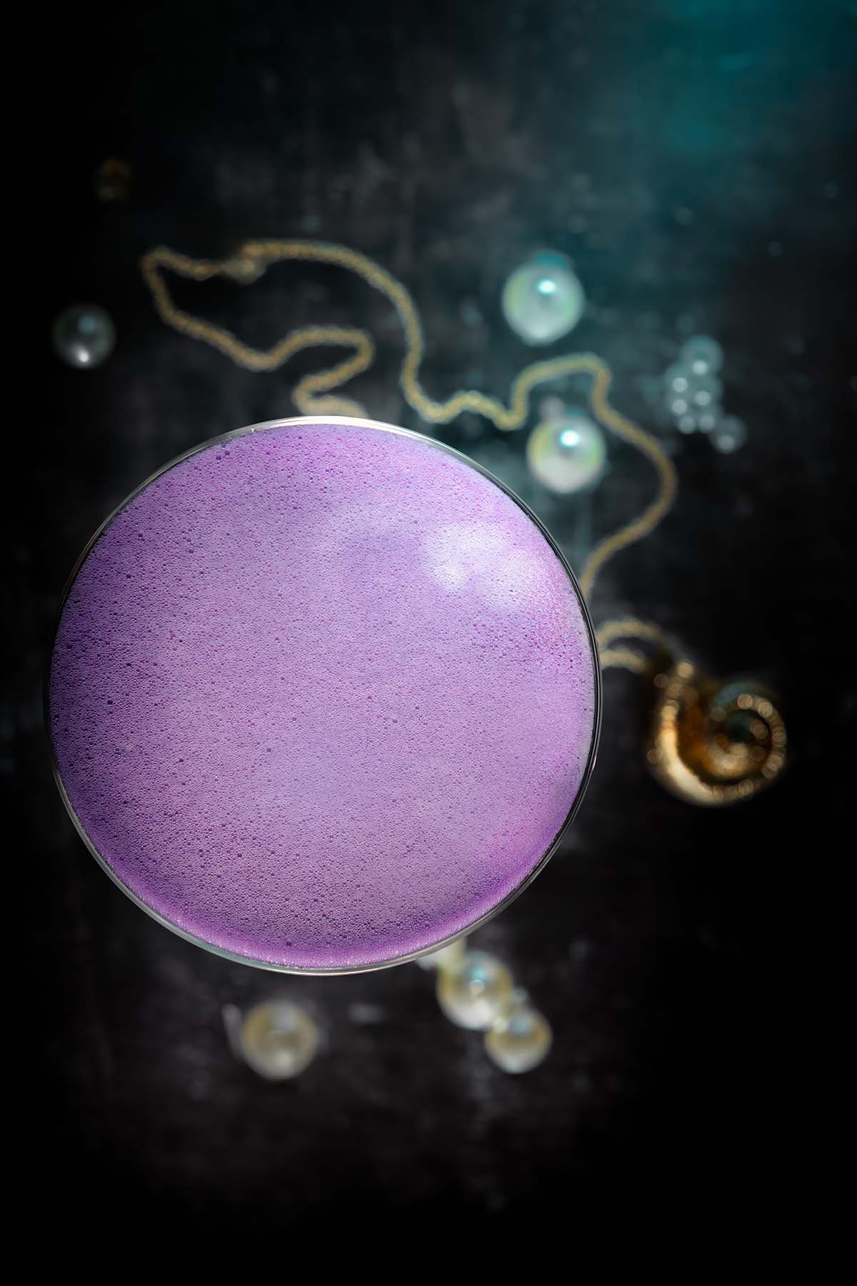 A top-down view of a lilac-foamed cocktail by Lou Carruthers, with gold jewellery and pearls creating an ethereal undersea atmosphere inspired by Ursula.