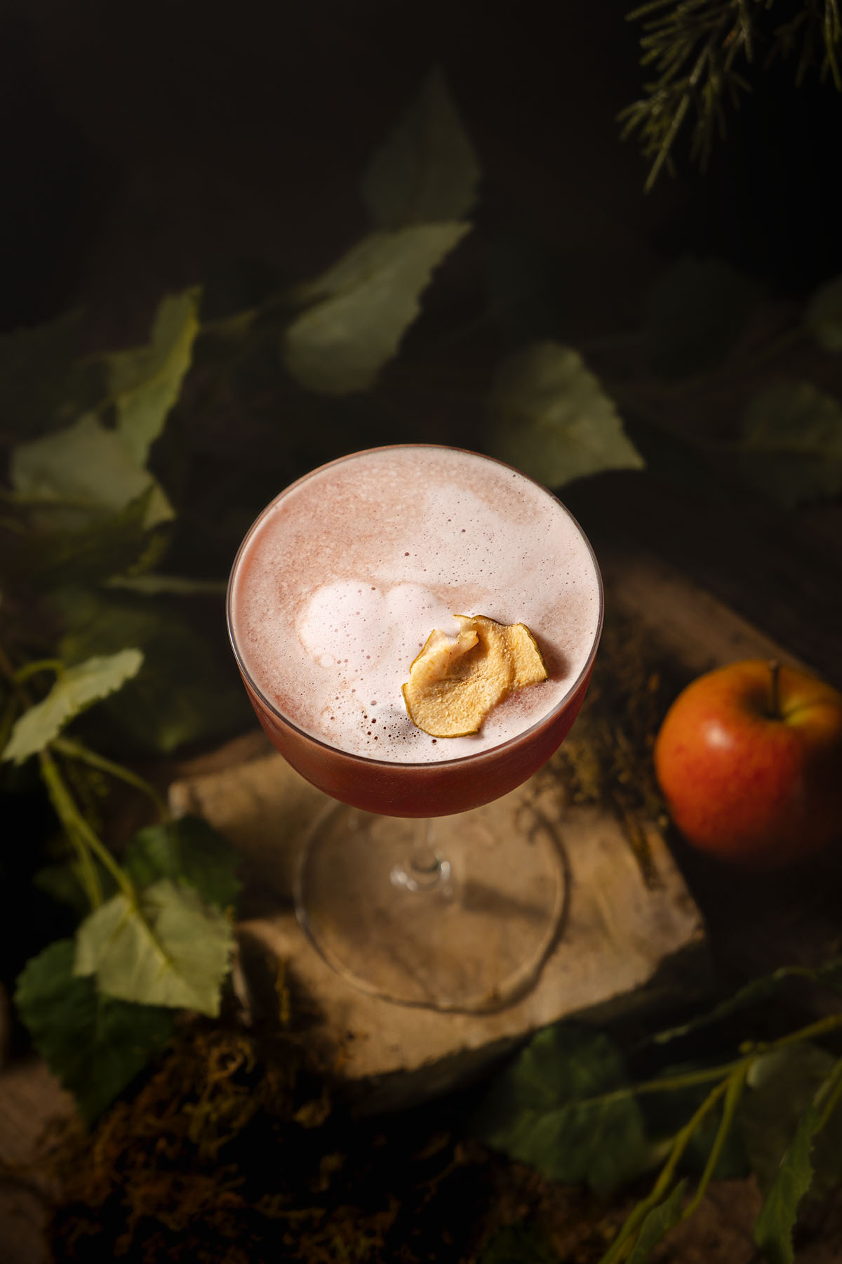 Overhead photograph of the Evil Queen mocktail by Lou Carruthers, showing a pale apple foam crown with a golden apple chip garnish. The drink sits on moss and stone with surrounding leaves, styled to evoke the dark forest of Snow White.