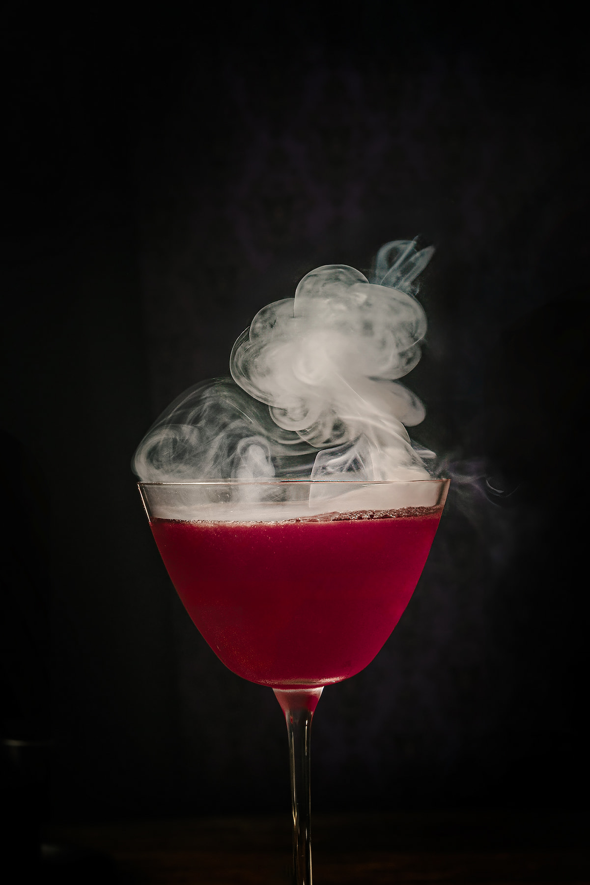 Close-up of a Halloween cocktail inspired by Maleficent, photographed by Lou Carruthers. The deep magenta drink swirls with delicate smoke rising from the glass, set against a dark, moody background. A dramatic and elegant gin-based cocktail perfect for spooky season styling and Halloween party inspiration.