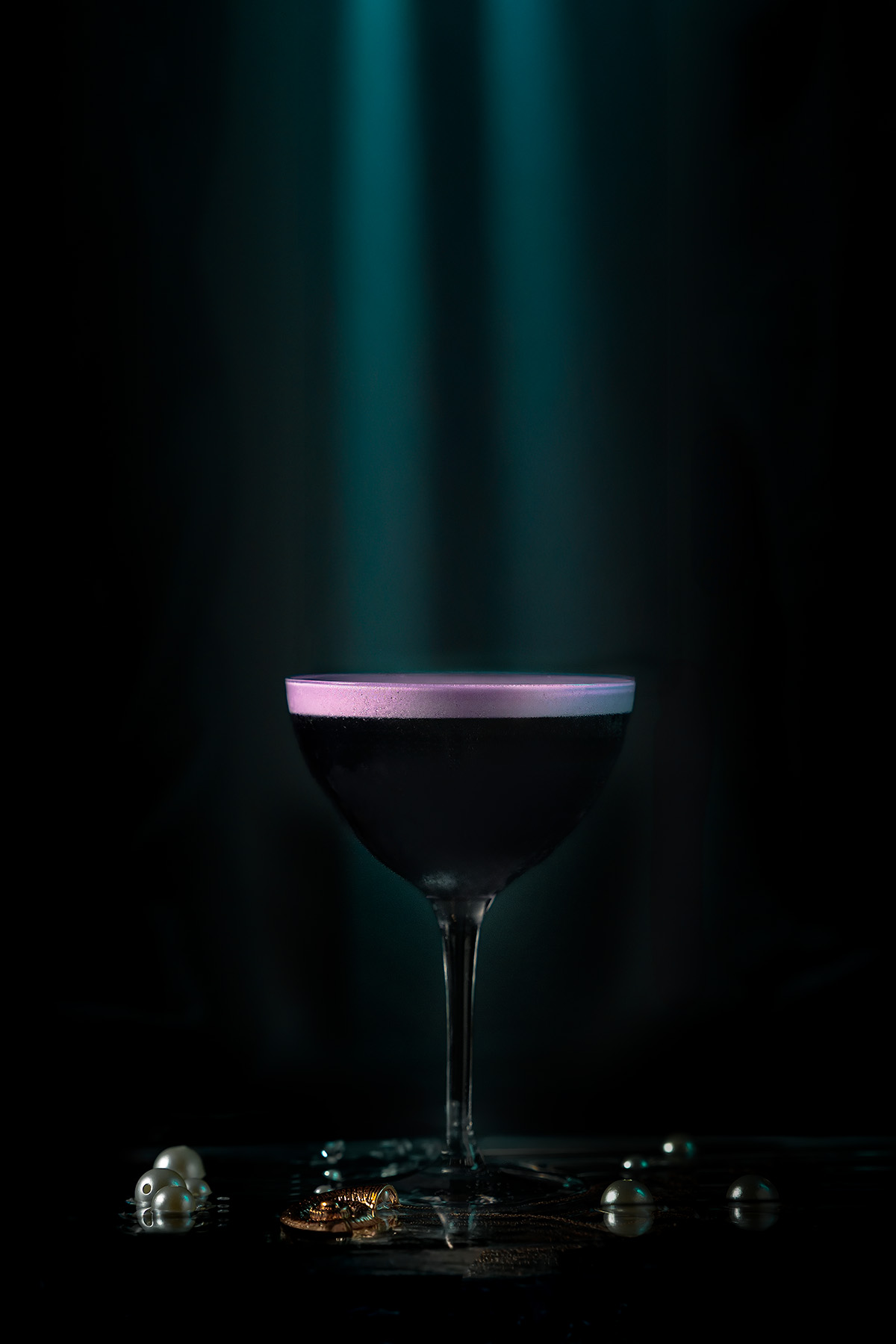 A dark cocktail under god-ray style teal light beams, with pearls and a gold seashell necklace glimmering below, photographed by Lou Carruthers as part of her Once Upon a Cocktail series.