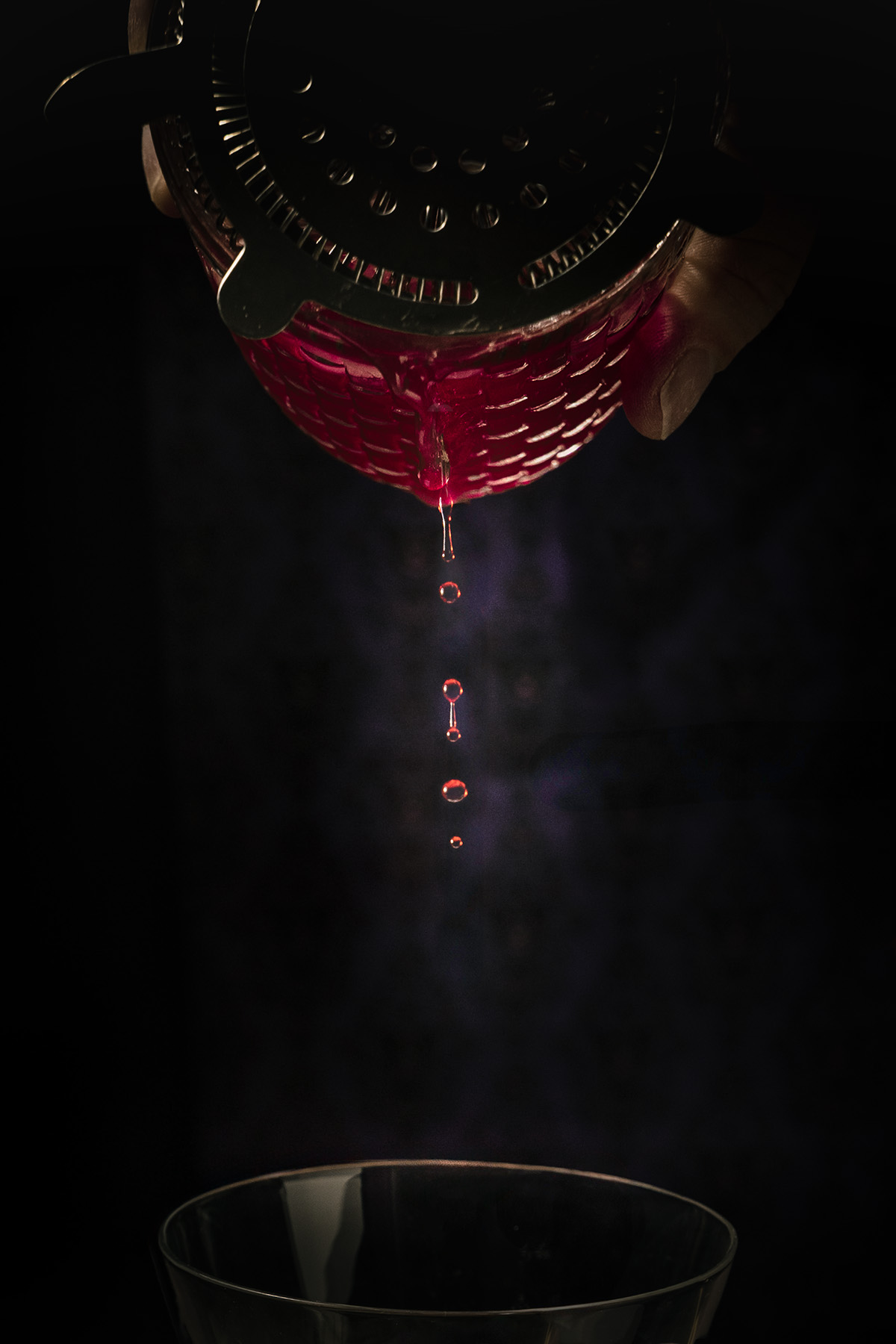 A close-up detail of the Maleficent Halloween cocktail by Lou Carruthers, showing droplets of rich magenta liquid falling from a strainer into a glass below. The moody lighting and dark background evoke the moment the spell begins — a subtle nod to Maleficent’s blood-red curse and gothic elegance.
